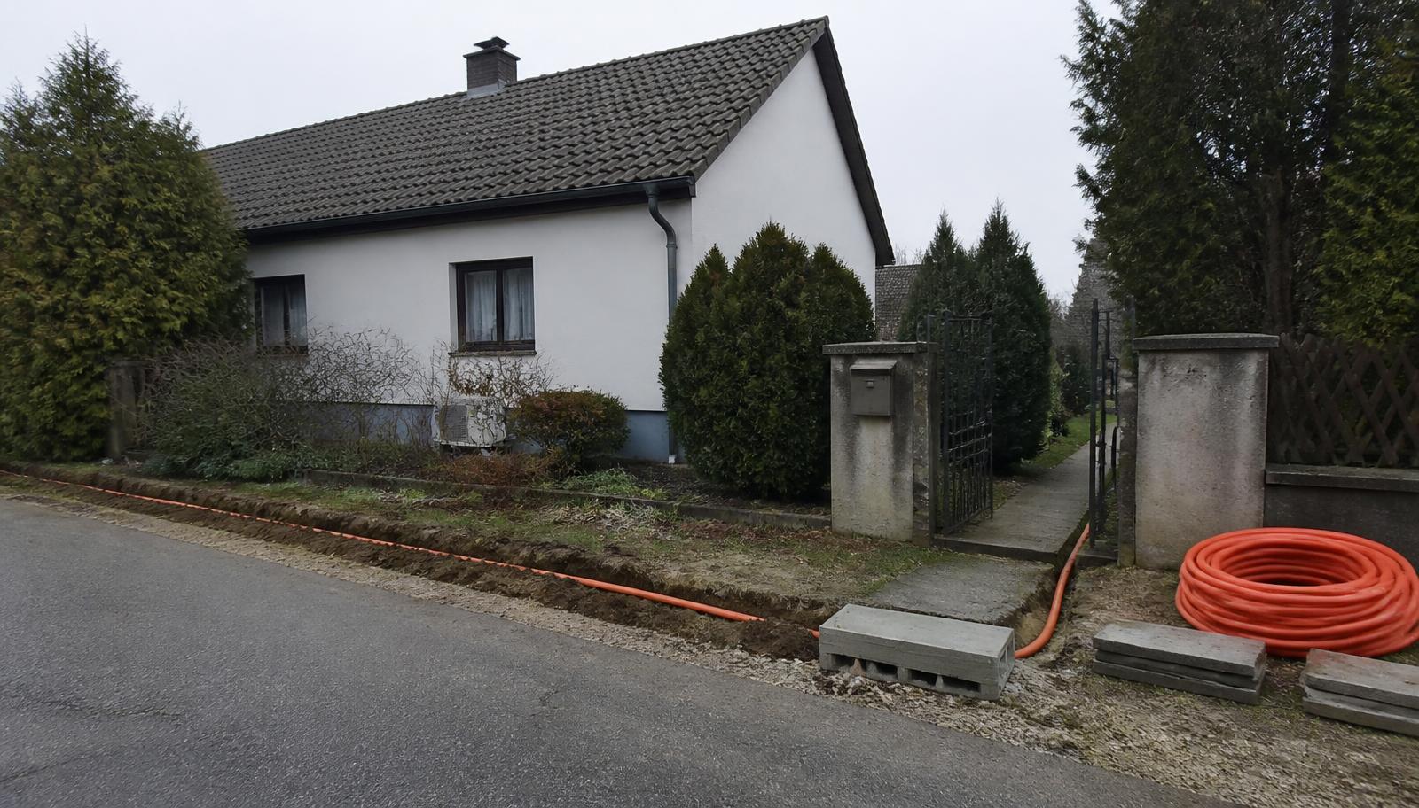 Offener Glasfasergraben mit orangem Leerrohr am Straßenrand vor einem österreichischen Einfamilienhaus